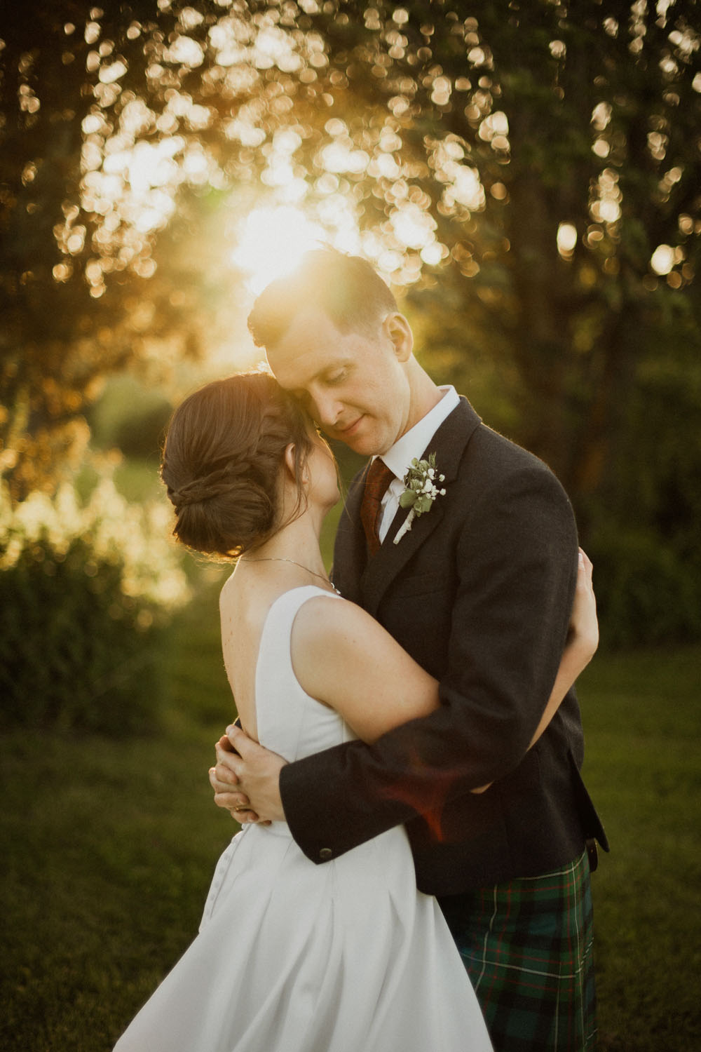 Porträttbild på brudpar som håller om varandra med låg sol i bakgrunden, Fotograf JD