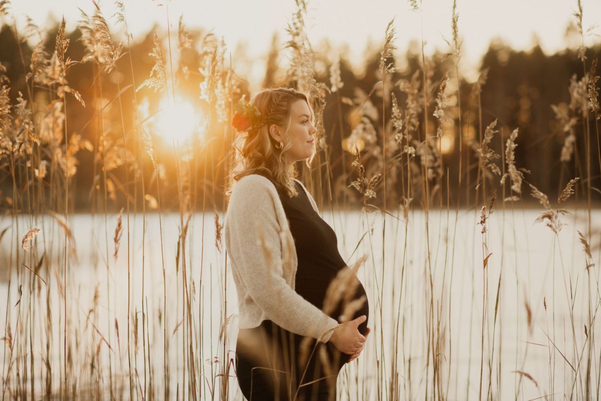 Gravid kvinna står vid vattnet omgiven av vass i låg solljus, Fotograf JD